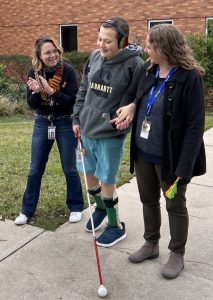 A young man in shorts and a sweatshirt, holds his cane and the arm of his PT while laughing with his mother on campus.