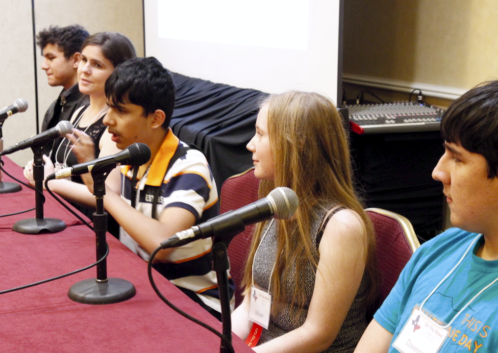 Five adolescents sit at a table with microphones during a panel discussion at a conference.