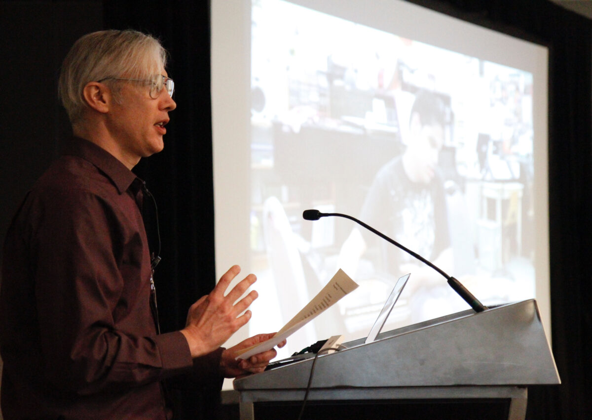 An adult dressed formally stands at a podium and speaks into a microphone next to a large screen depicting an adolescent.