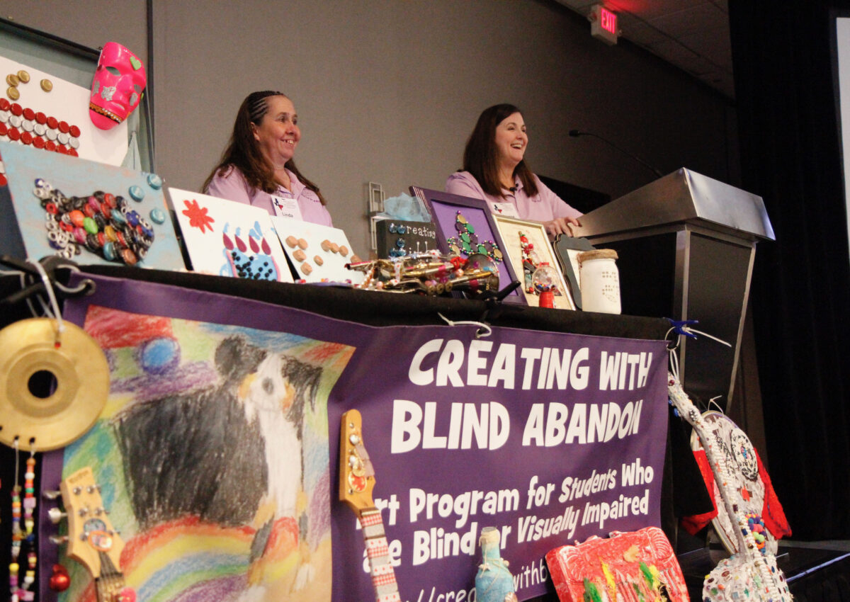 Two adults stand at a podium and behind a table decorated with various types of tactile and visual art while presenting at a conference.