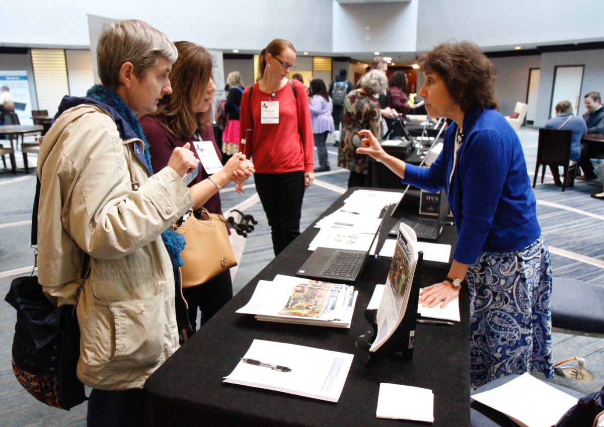 In a hotel foyer set up with information booths, an adult standing behind a table speaks to two interested adults. On the table are paper publications of TX SenseAbilities.