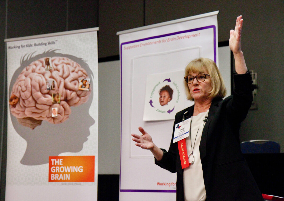 An adult stands on stage and presents at a conference with two large graphics behind her, 