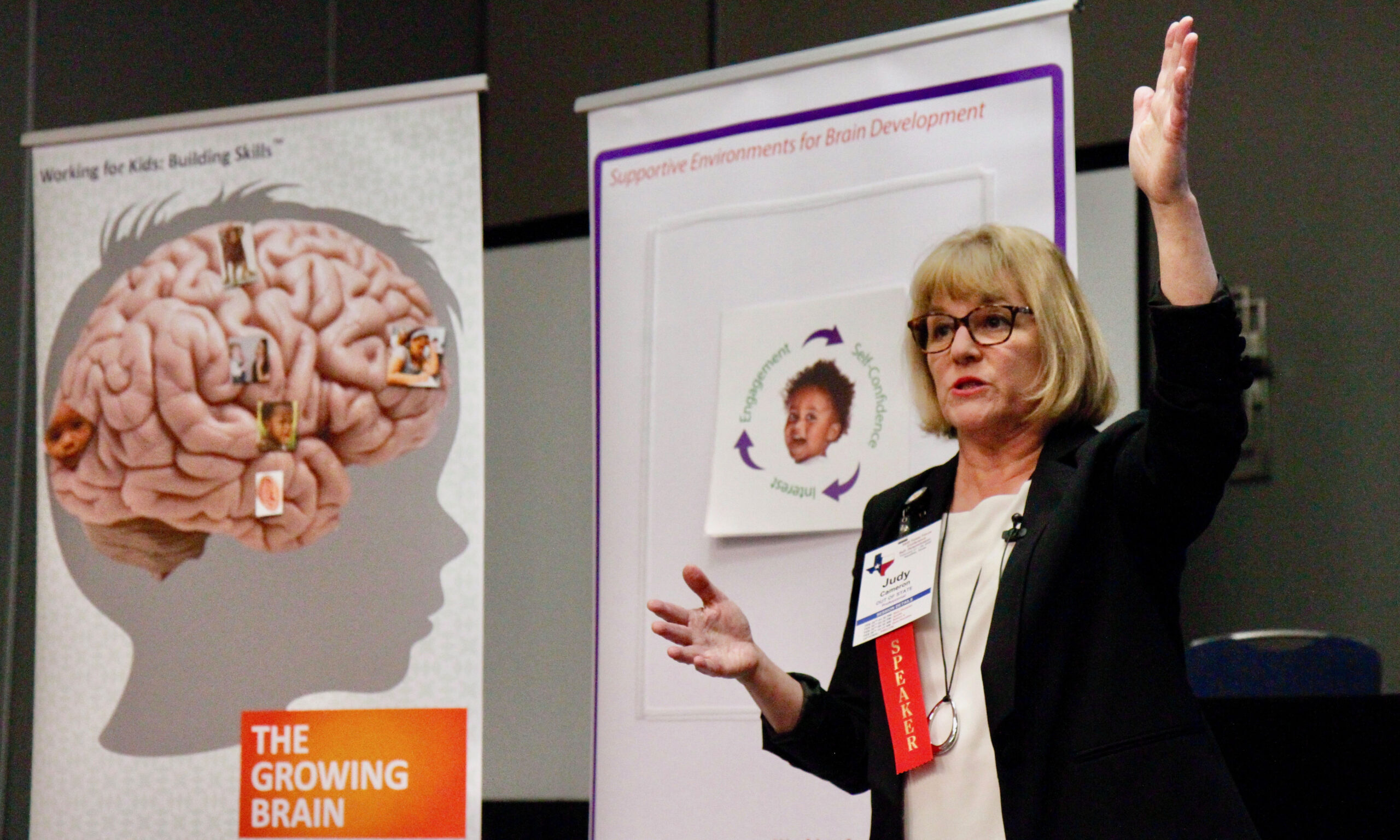 An adult stands on stage and presents at a conference with two large graphics behind her, "Working with Kids: Building Skills" and "Supporting Environments for Brain Development."
