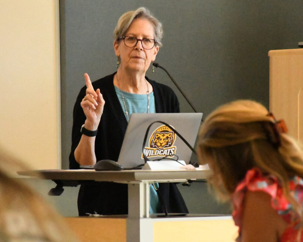 An adults stands in conference room at a podium with a laptop with TSBVI Wildcats sticker. Her finger is up and she talks to rows of adults.
