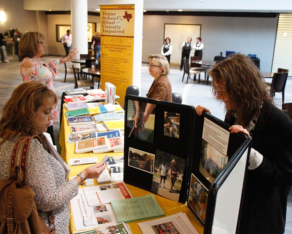 Conference attendees visit with TSBVI employees at a table with resources, curriculum, and a trifold board. In the background, other attendees and staff visit and wander a conference lobby.