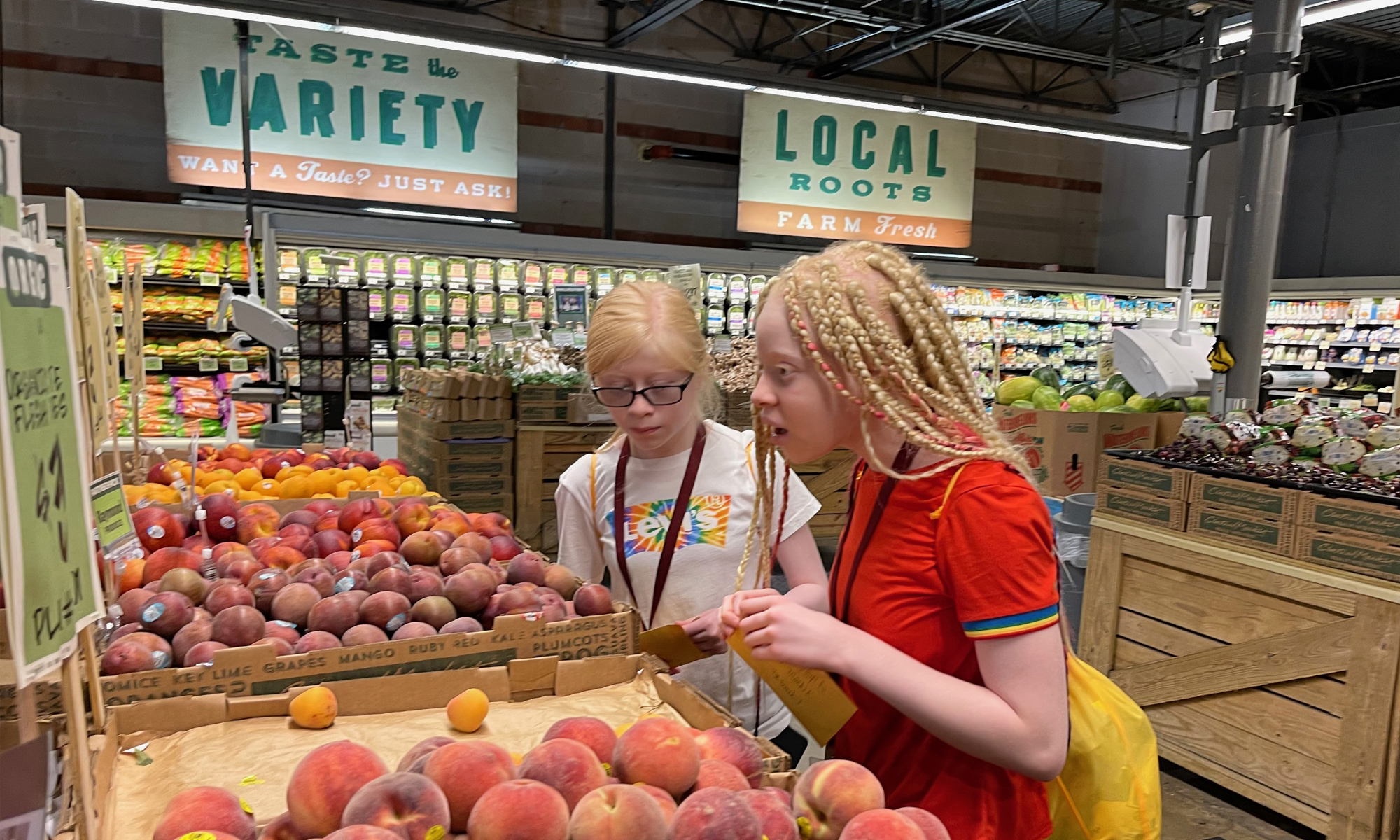 Two adolescents stand in front of bins of peaches in the produce department of a large grocery store.