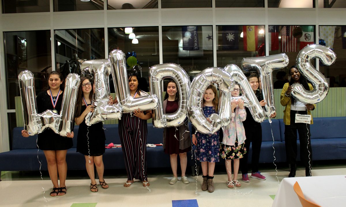 Cheerleaders and coach in semi-formal wear holding balloons that spell out 