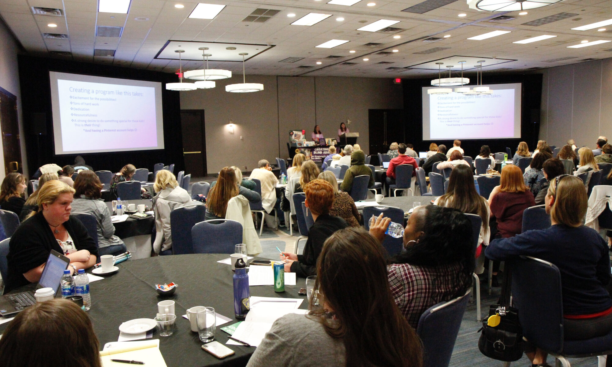 Conference attendees sit at round tables in a ballroom to listen to a presenter.