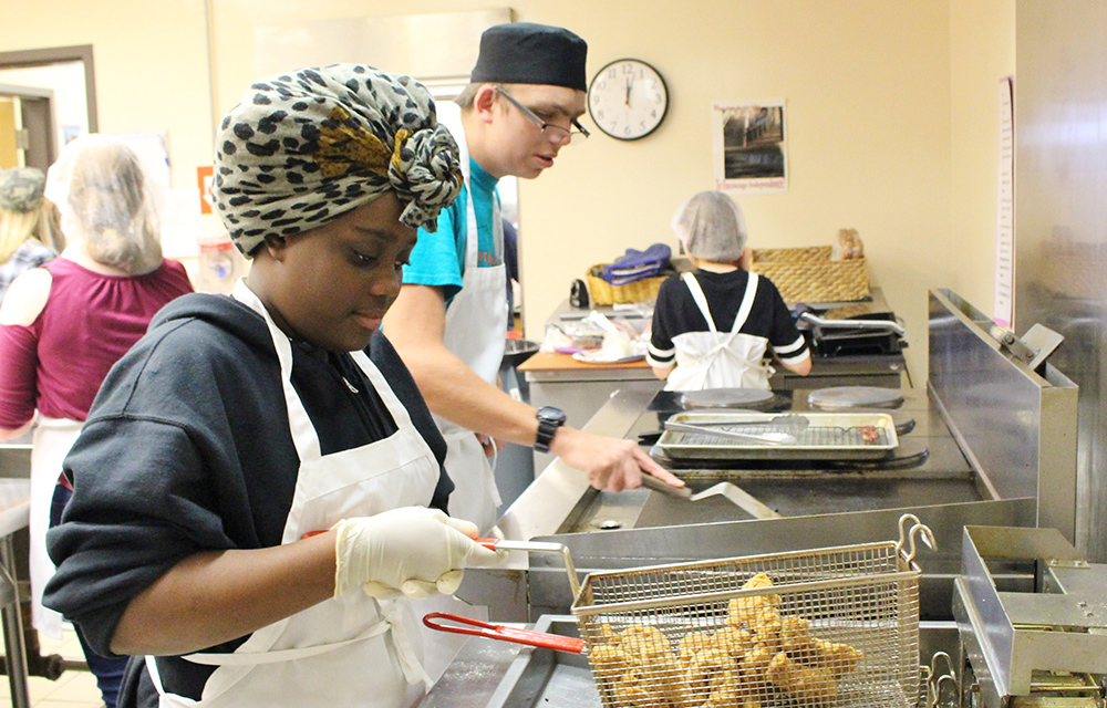 Student working in a kitchen frying food with another student in background