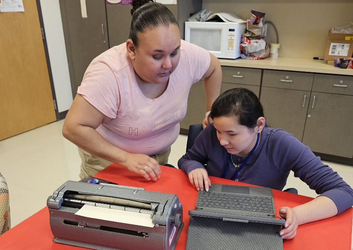 An adult instructor with a visual impairment instructs a student using a laptop and a Perkins Brailler.