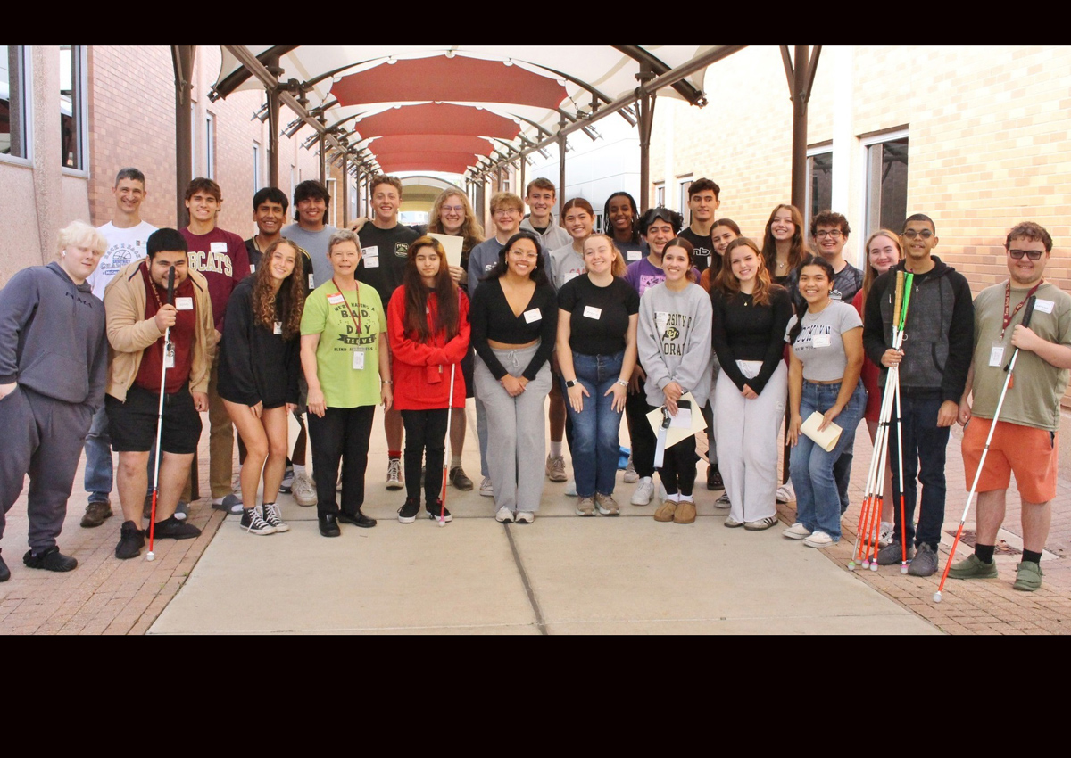 A large group of adults and adolescents, some with white canes, stand under a covered walkway outdoors.