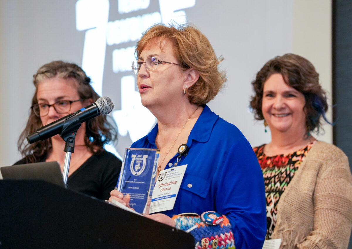 An adult at a podium holds a glass award while two adults smile behind her.