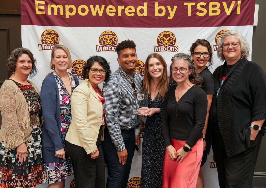 Eight adults stand in front of a backdrop that says "Empowered by TSBVI." One holds a glass award.