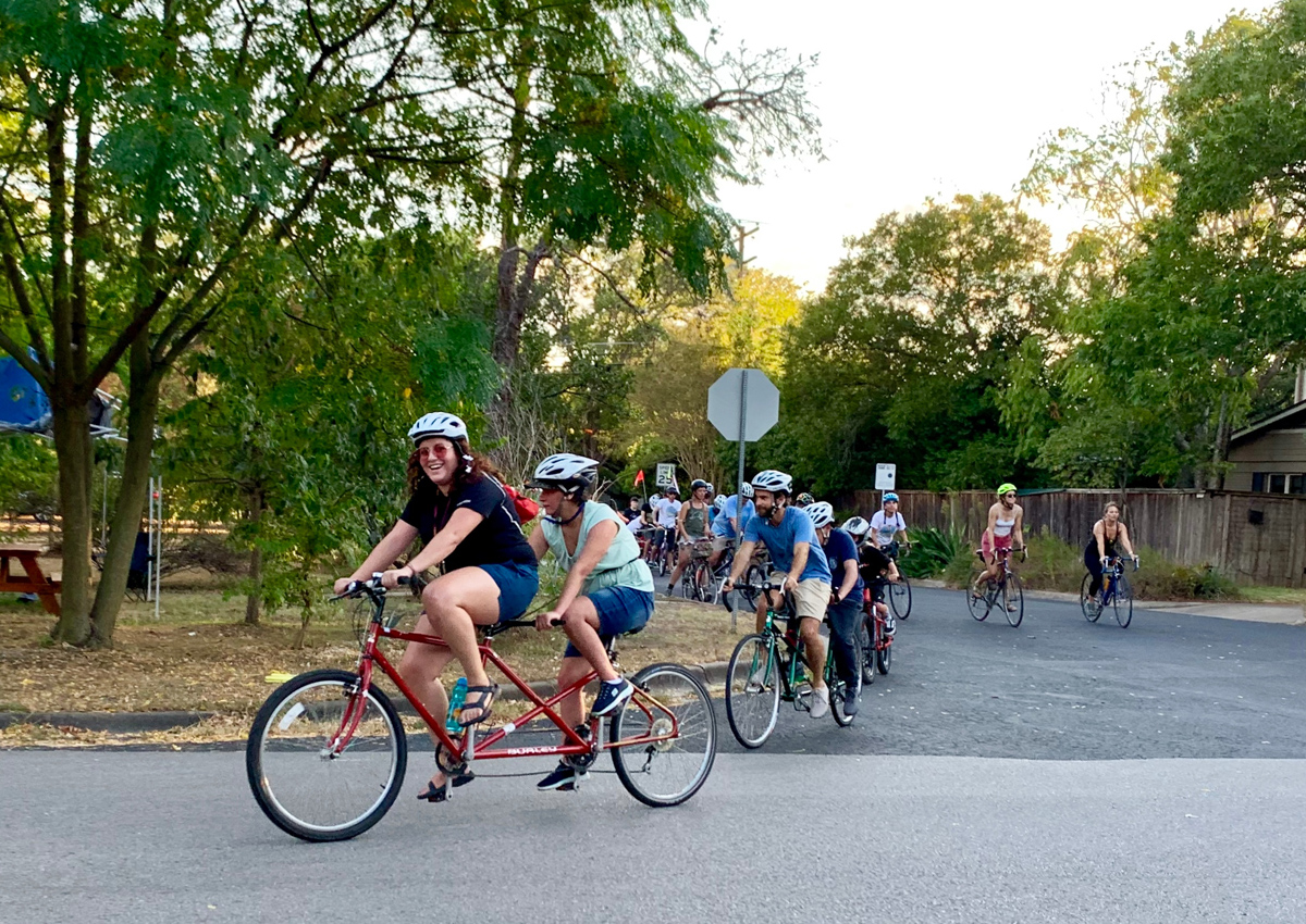 Un grupo de adultos y adolescentes en bicicletas tándem y estándar recorren una calle residencial.