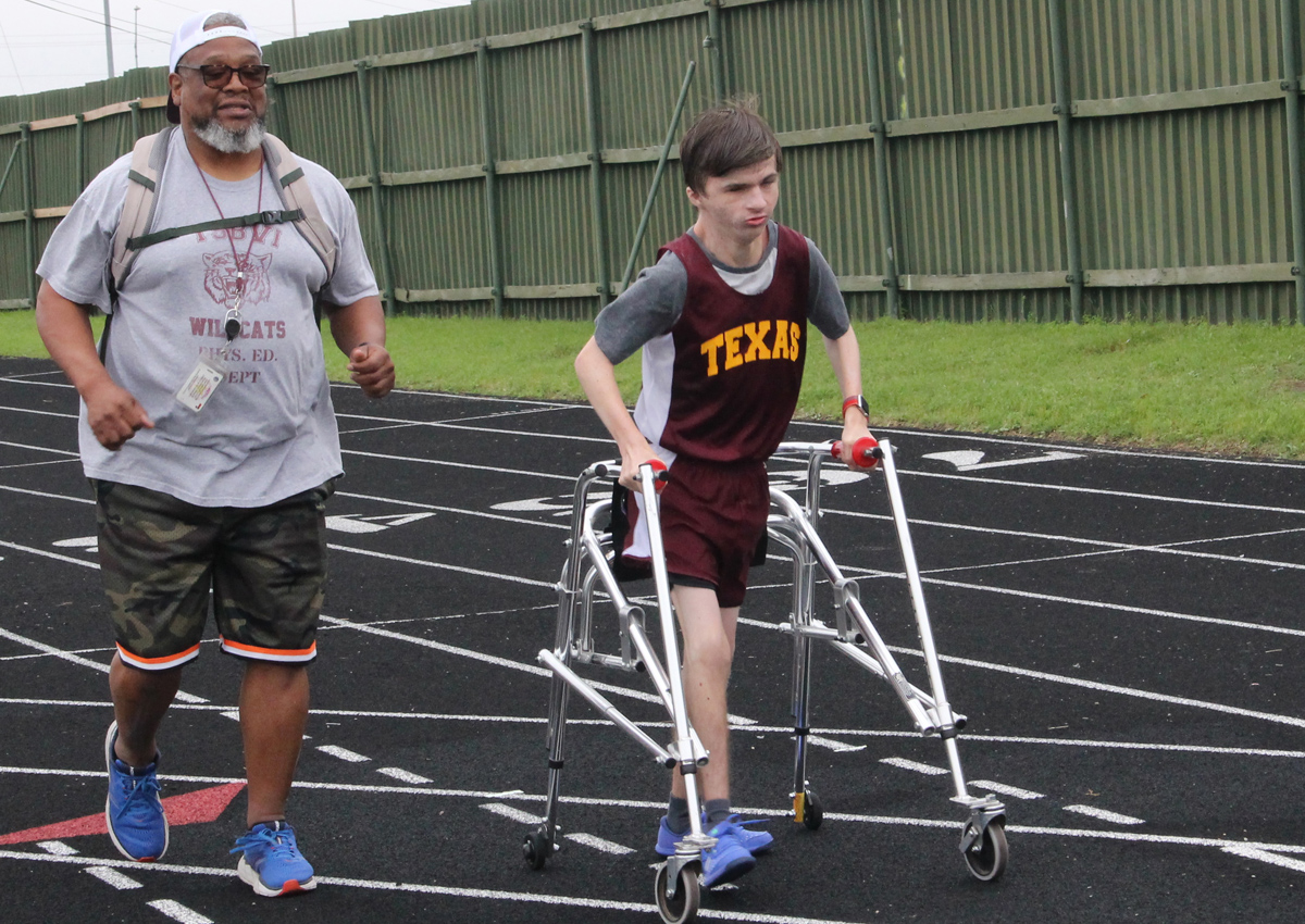 Un adolescente que utiliza un andador corre por una pista con un adulto que corre a su lado.