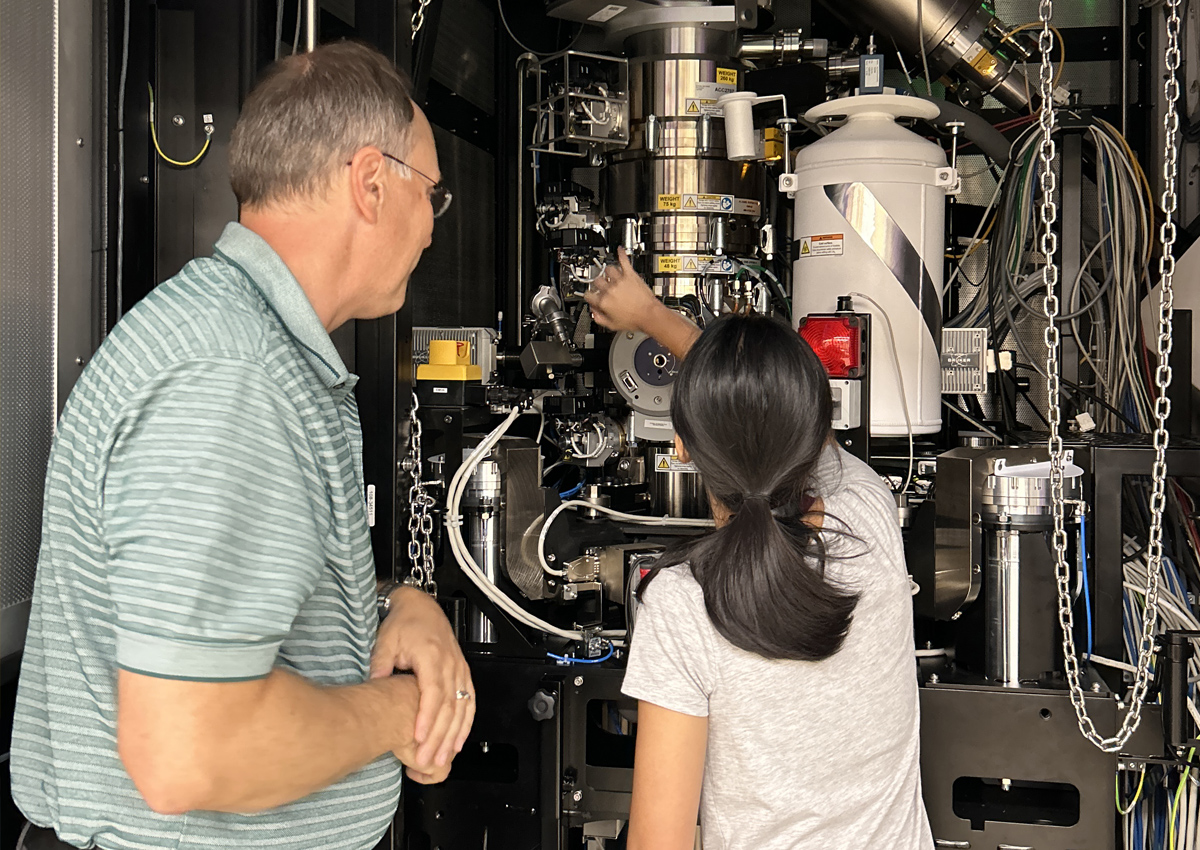 Una adolescente toca un microscopio electrónico de transmisión grande que llega hasta el suelo mientras un adulto está de pie a su lado.