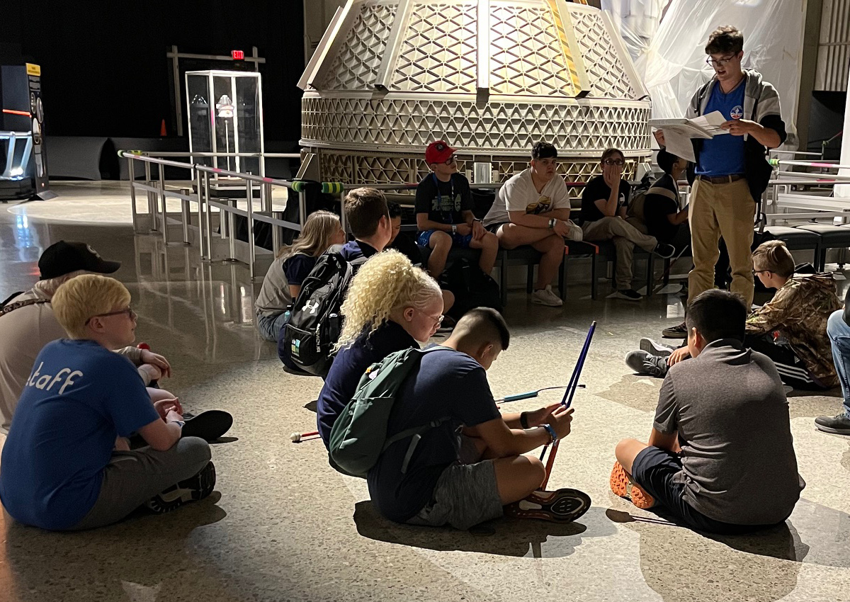 Un grupo grande de adolescentes se sientan en el suelo y en sillas mientras un adulto les lee en un museo espacial.