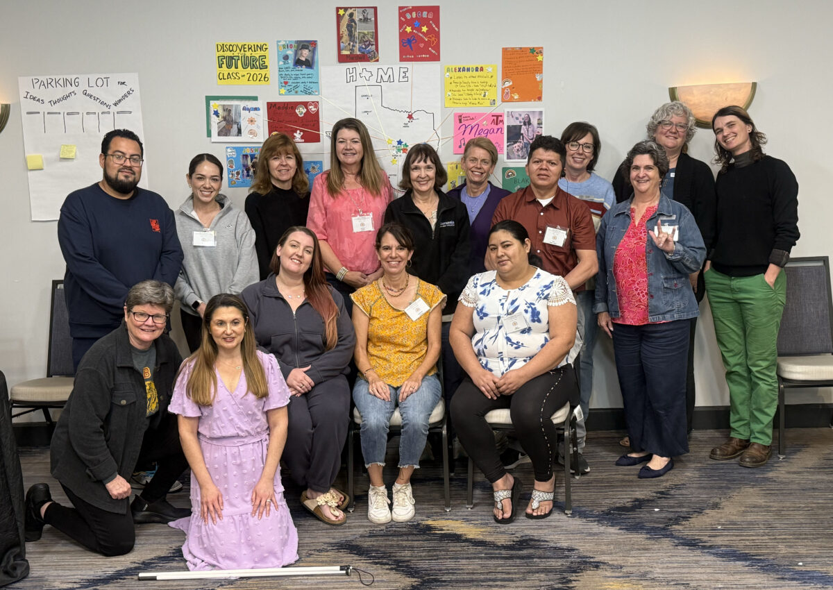 A large group of adults stand and sit in a conference room in front of artistic displays representing their children and the word 