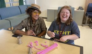 An adult smiles and an adolescent laughs while seated at a table with toilet paper rolls cut with little slits and pink pipe cleaners in curlicues.