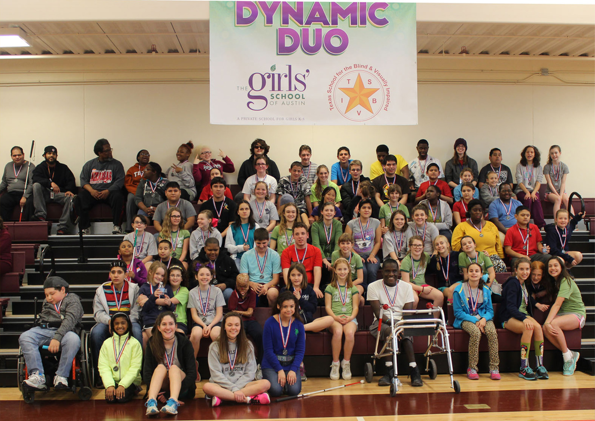 A large group of adolescents and adults fill risers in a gym with a banner reading 