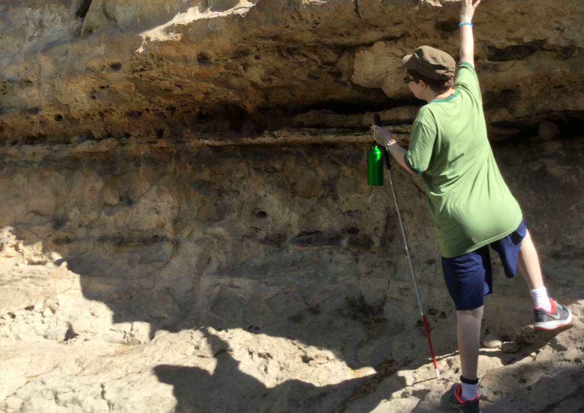 An adolescent with a white cane feels the limestone rock hanging overhead while on a hike.