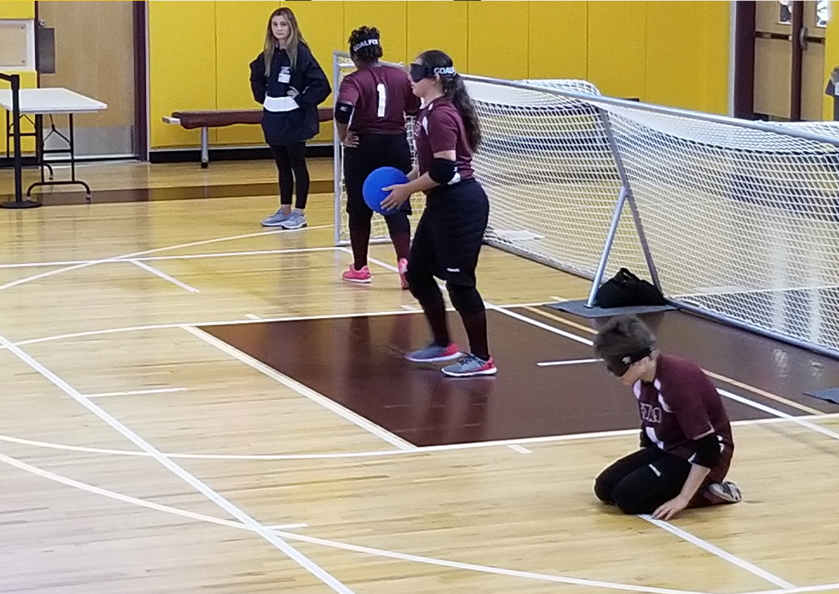 Texas girls team playing goalball in the TSBVI gym with a coach watching on the sideline.