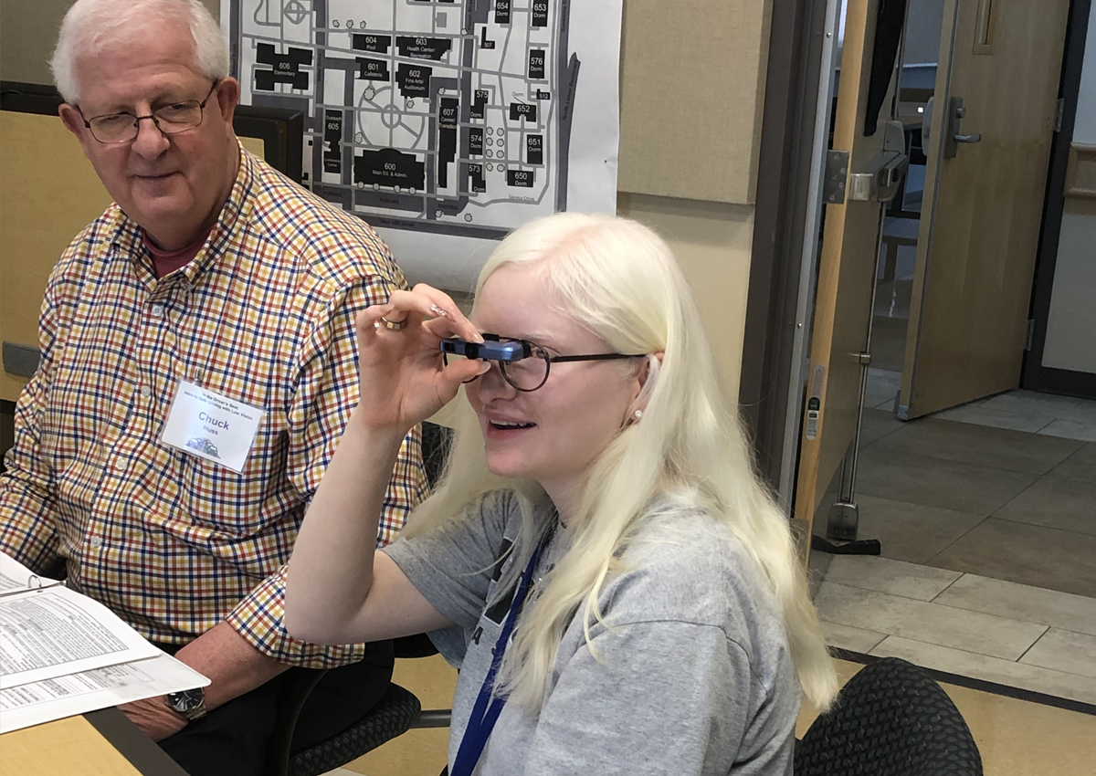 An adolescent looks through bioptic glasses while an adult sits next to her.