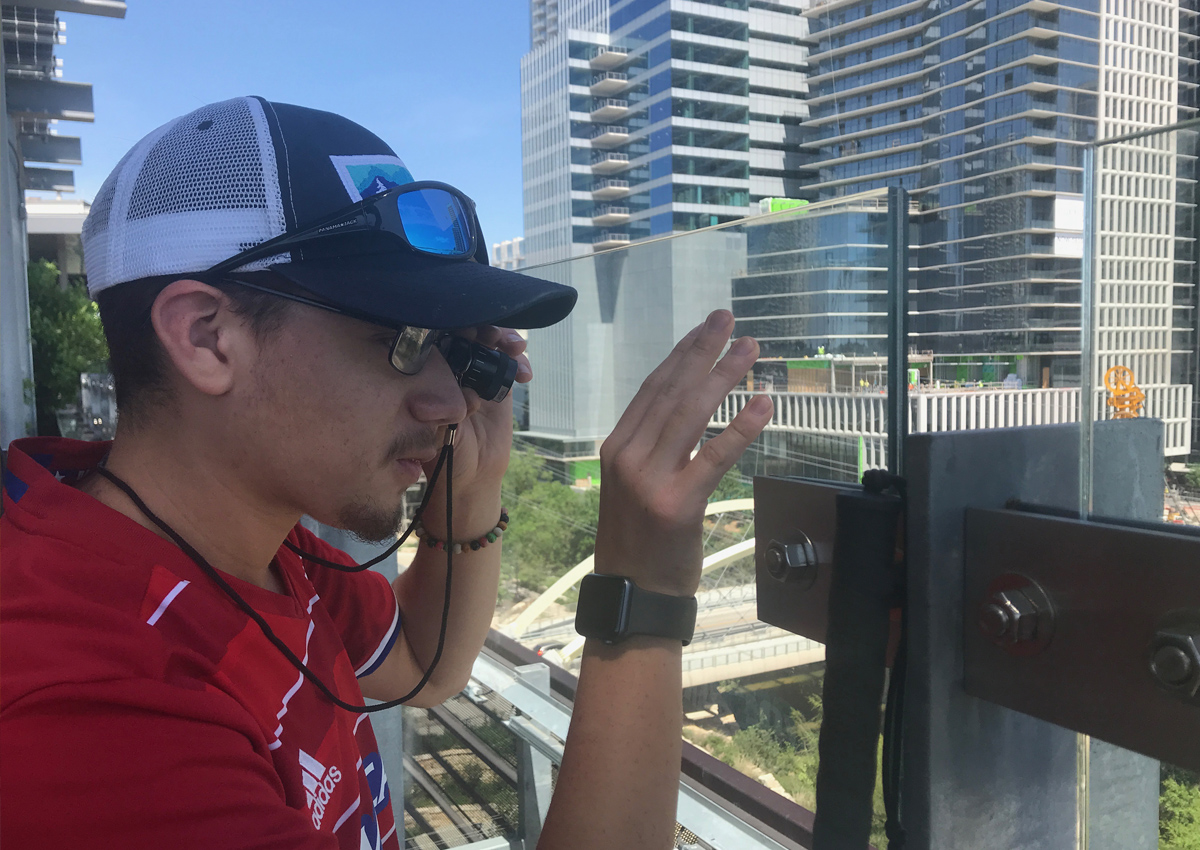 An adolescent stands on a downtown balcony with his hand on the glass partition and uses a telescope to look at the skyscrapers and cityscape below.