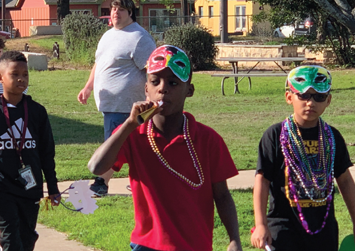 Five students walk outside wearing beaded necklaces and homemade, painted face masks while blowing noisemakers.