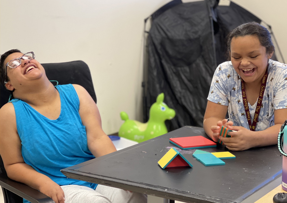 Two adolescents sit at a table in a classroom with magnetic tiles on the table. They both laugh hysterically.