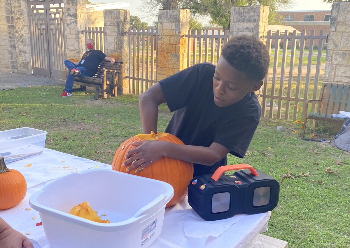 An adolescent reaches into a large pumpkin on a picnic table outside. A bin and a speaker are on the table.