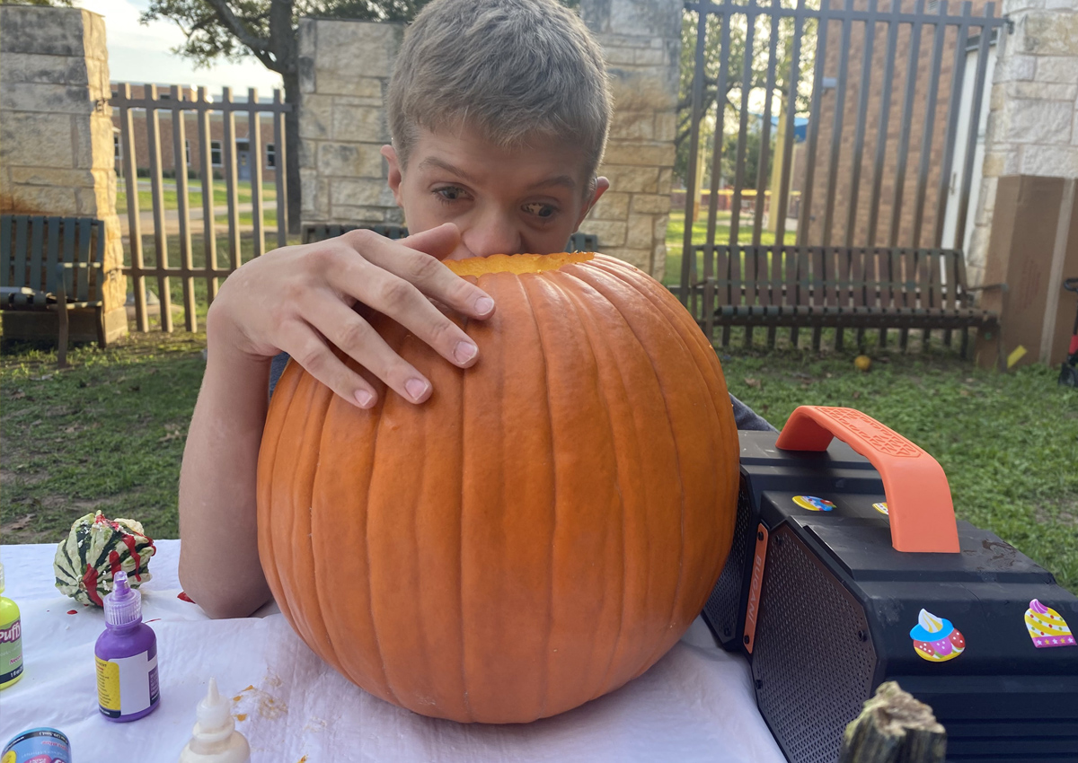 An adolescent holds his hand on a hollow pumpkin with a boom box resting next to it on a table outside.