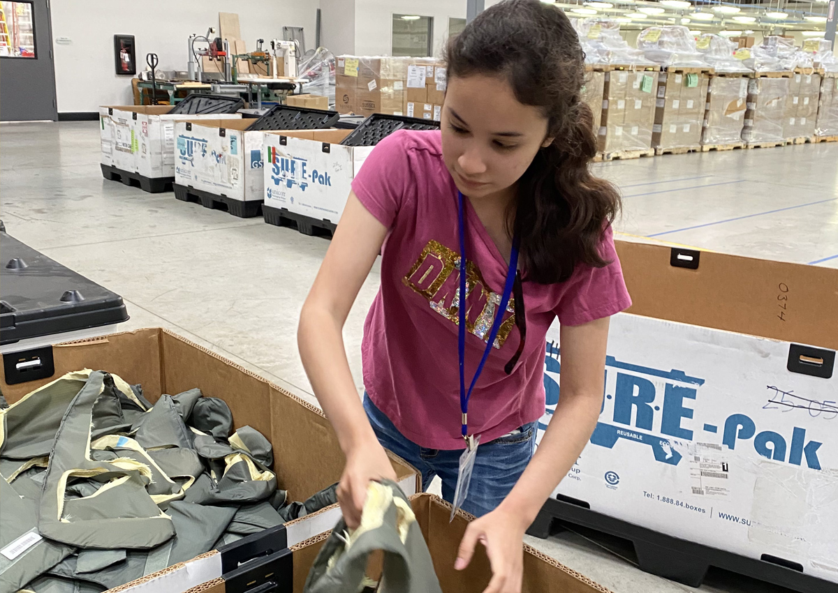 An adolescent reaches into a large box of refurbished military gear and pulls out a piece of equipment in a large warehouse with many bins and crates.
