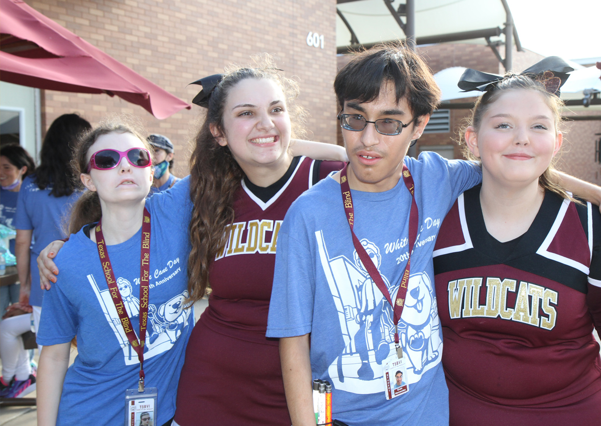 Two adolescents wear White Cane Day 2022 shirts and two adolescents wear cheerleading outfits outside during White Cane Day 2022.