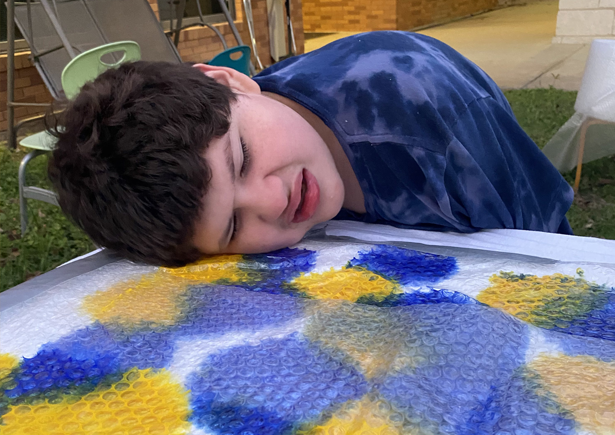 An adolescent seated at a picnic table outside rests his head on a painting of bright blue and yellow dots under a sheet of bubble wrap.