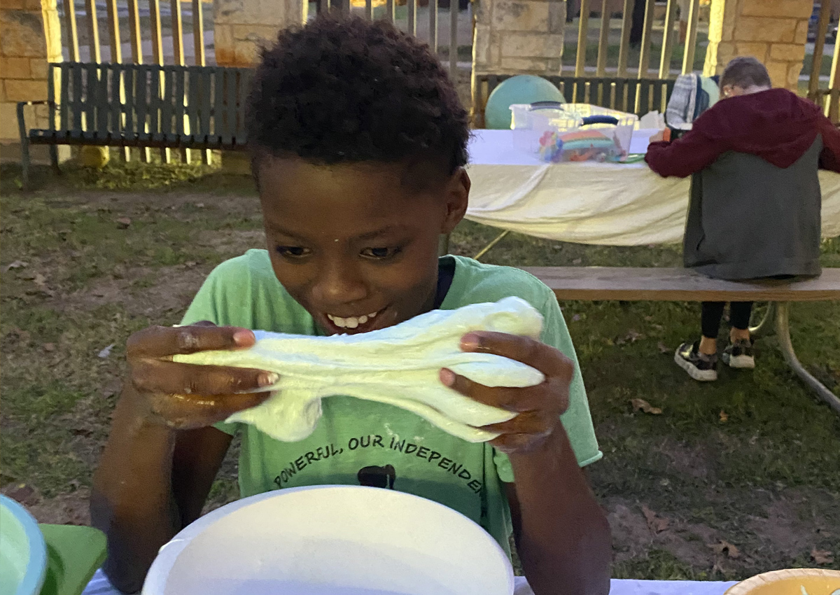 An adolescent seated at a picnic table outside stretches a wad of light green marshmallow slime.