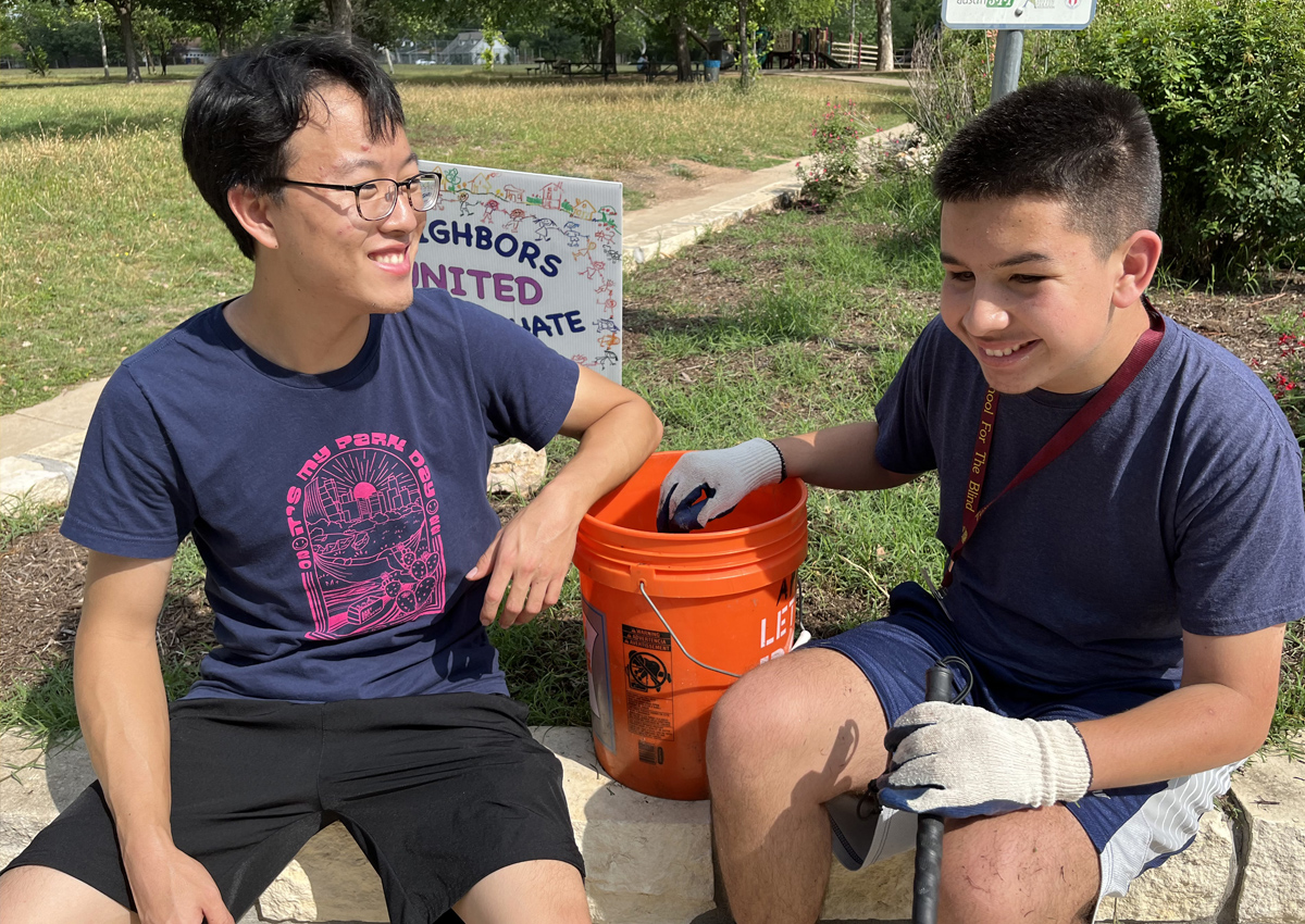 An adolescent wearing gloves pulls weeds into a bucket at a city park. He is seated next to a volunteer wearing a 