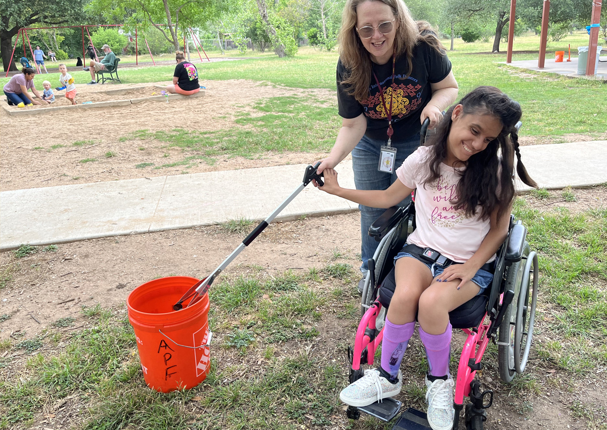 An adolescent in a wheelchair and adult hold onto a long trash picker upper and use it to place a piece of trash into a bucket at a park.