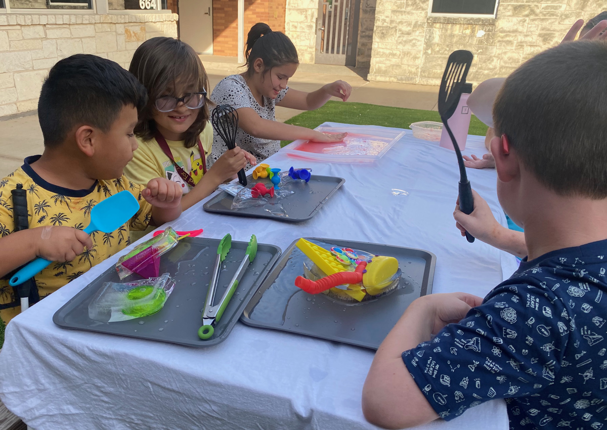 Four adolescents sit at a picnic table outdoors with trays of toys in blocks of ice and flavored ice.