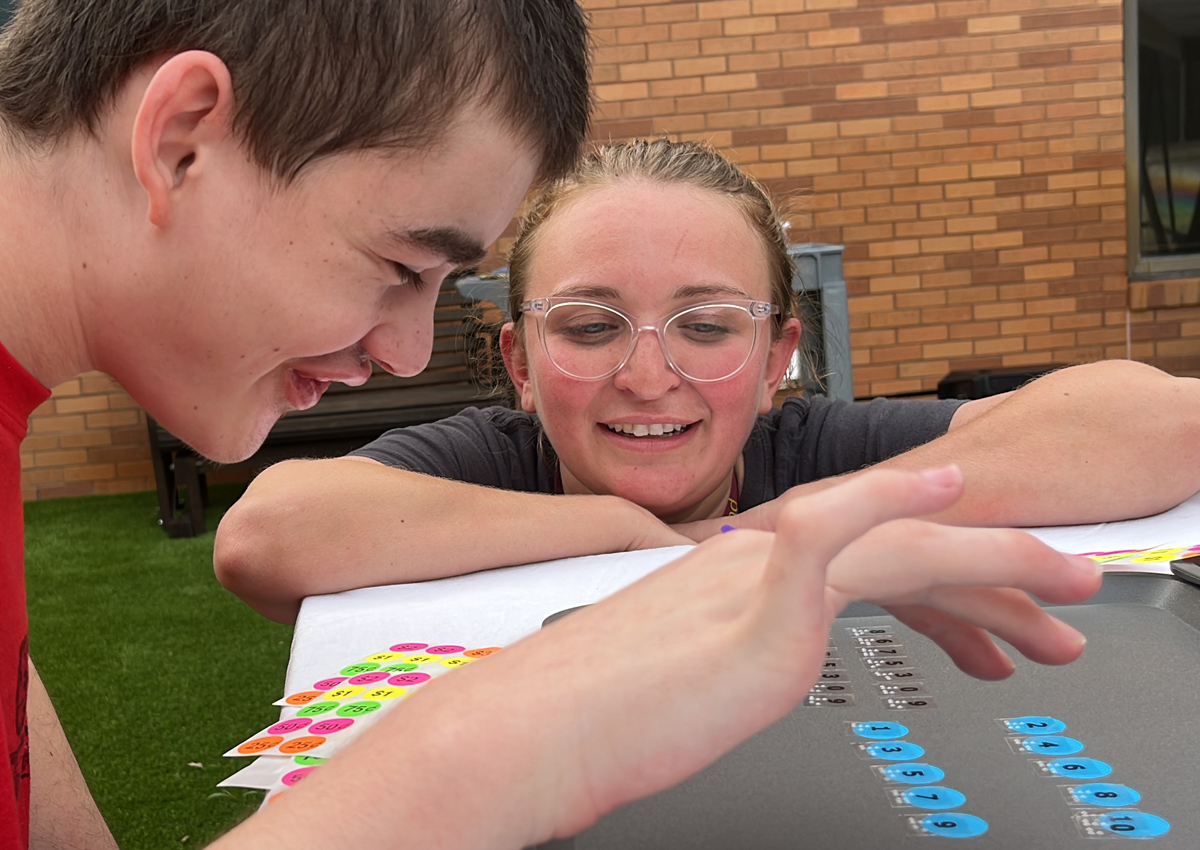 An adolescent seated at a picnic table outdoors reads number stickers with braille labels while an adult smiles next to him.