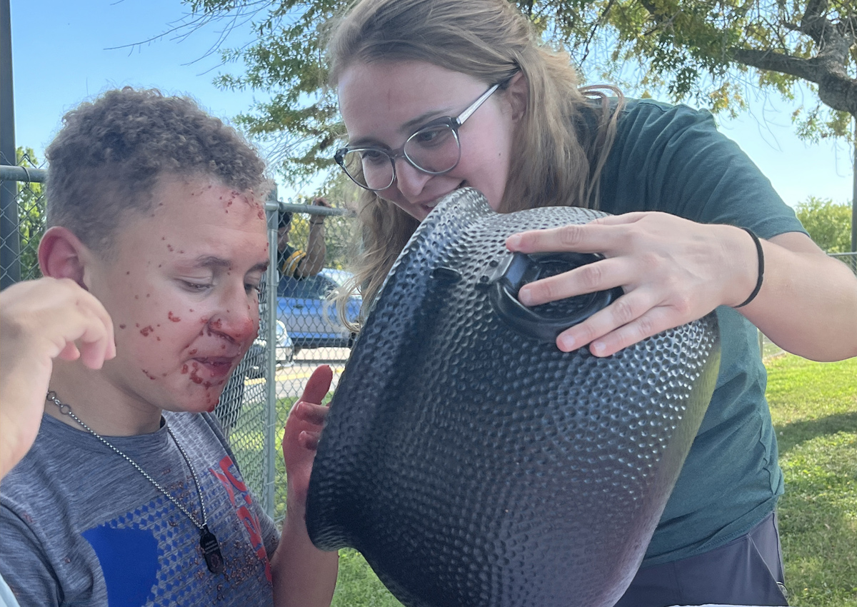 An adolescent with face and hands covered in red slime sits at a picnic table outside while an adult holds a large plastic cauldron to his mouth.
