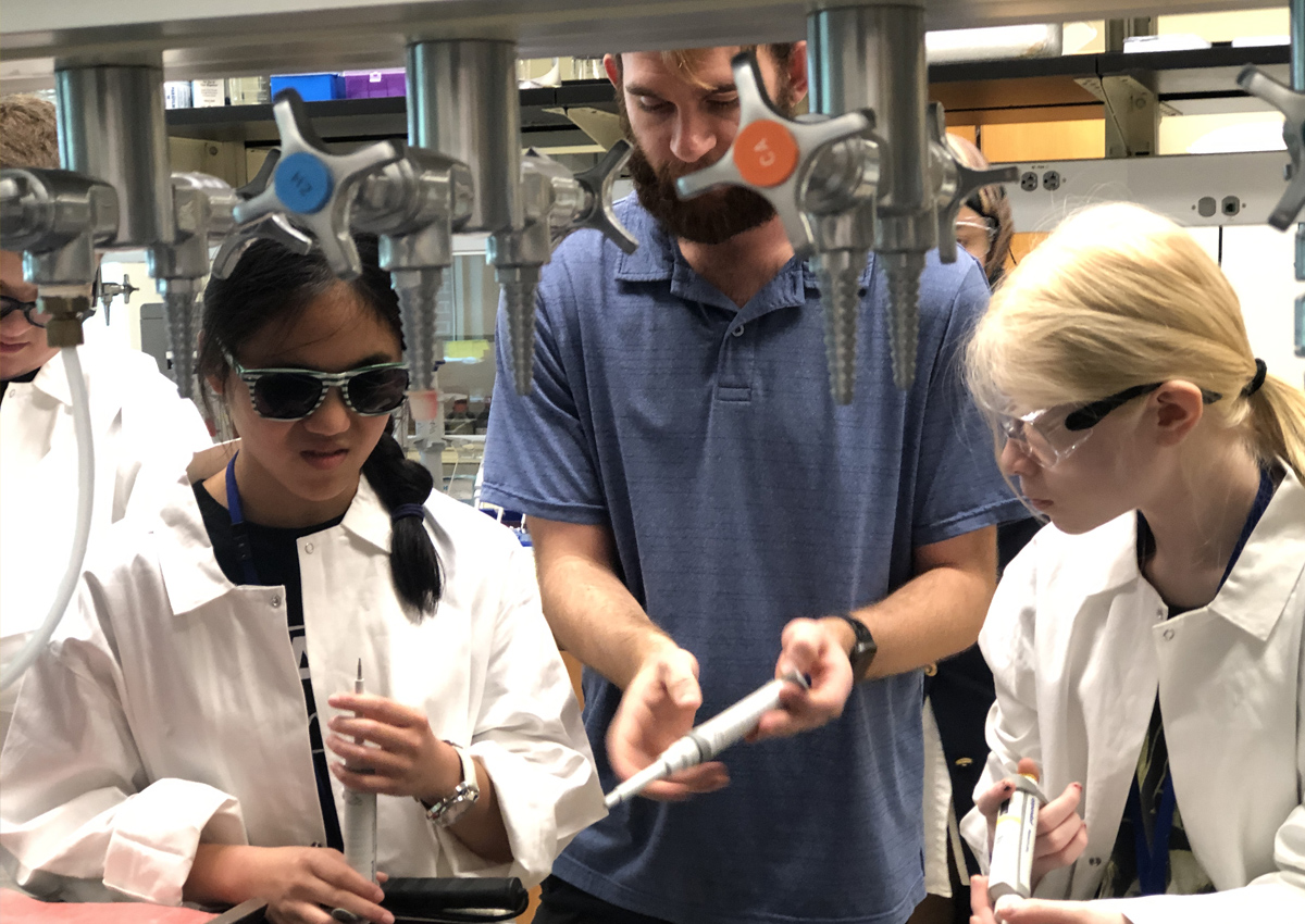 Twi adolescents in lab coats and protective eyewear hold pipettes and receive instruction from an adult holding a pipette in a chemistry lab.