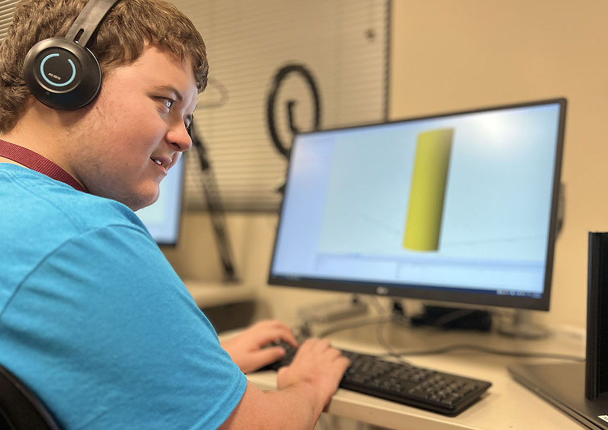 An adolescent wearing headphones sits at a computer with an image of a computer-generated large yellow cylinder on the screen.