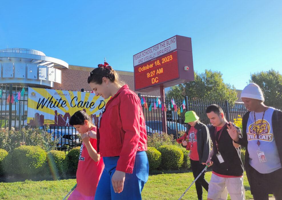 Adolescents and adults using tactile sign language march in front of the TSBVI Main Building 600 during the 2023 White Cane Day march.