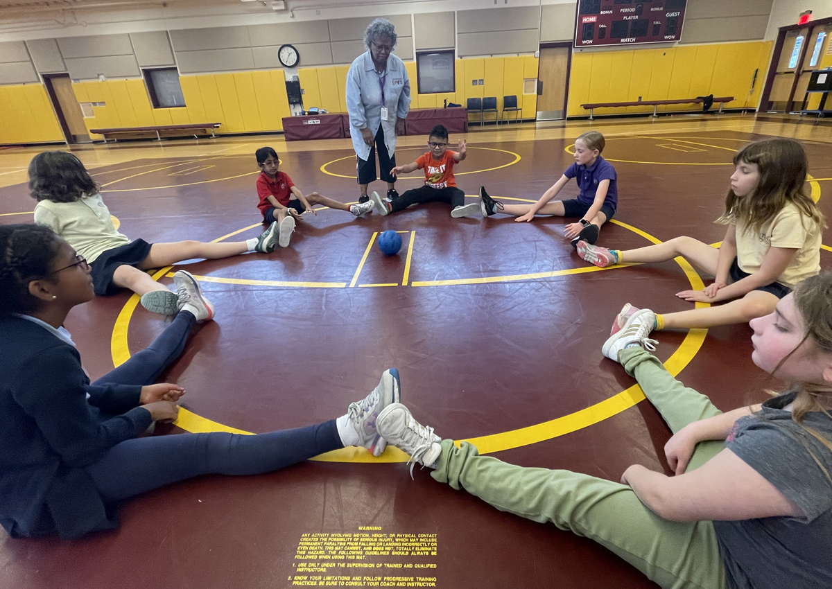 A large group of adolescents form a large circle on a gym floor and roll a ball with sound to each other while an adult stands around them.