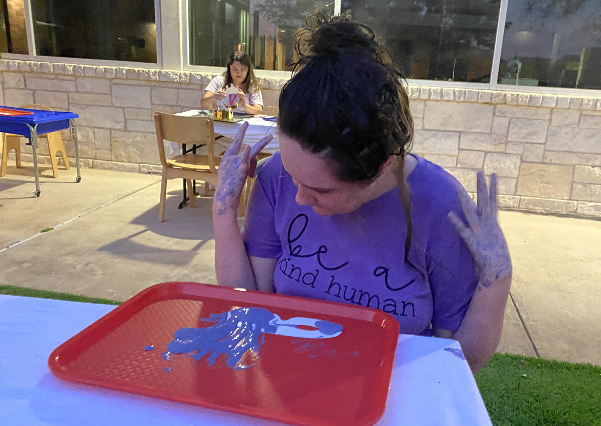 An adolescent sits at a picnic table outdoors with a Mardi Gras mask in a tray with purple paint on the table.
