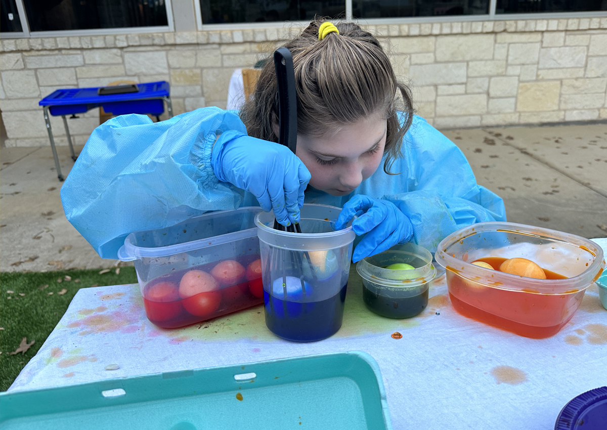 An adolescent in a smock seated at a picnic table dips eggs into colored water with egg dying supplies on the table.
