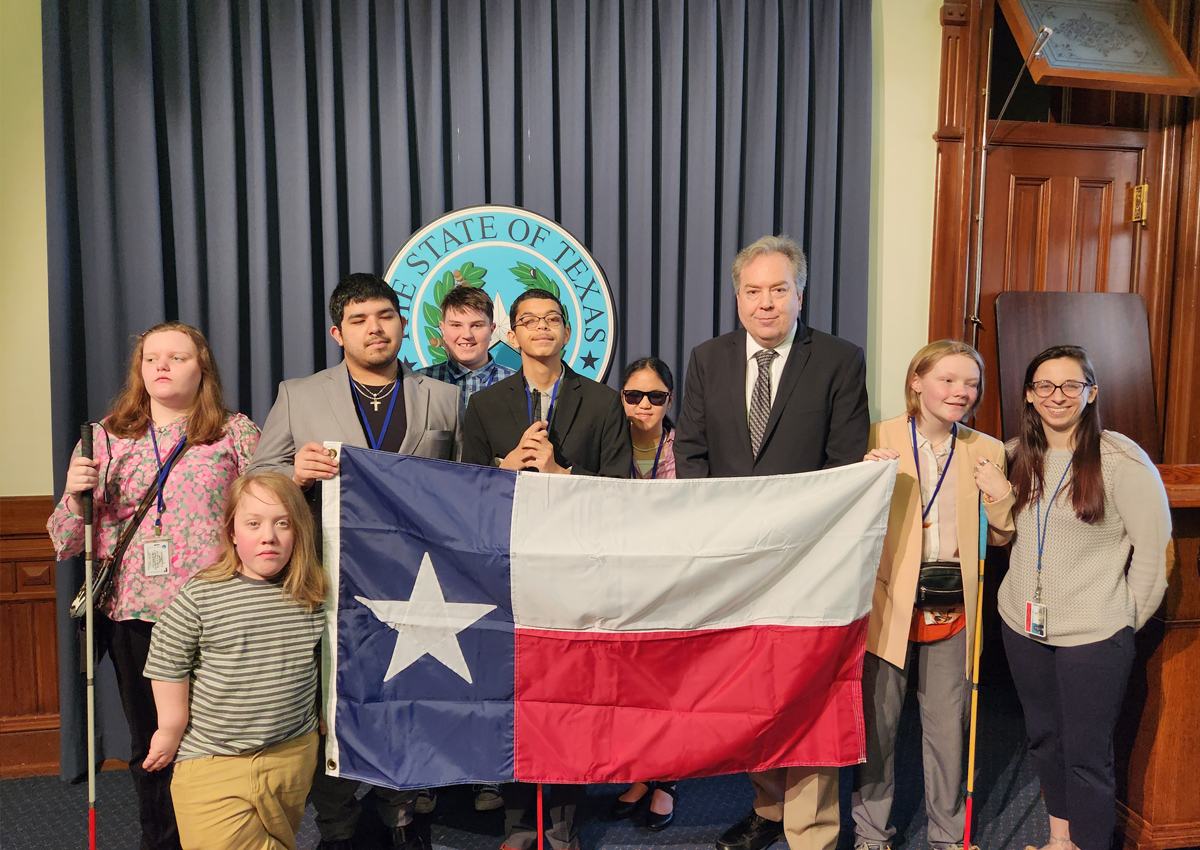 A group of adolescents in formal attire stand with Ron Lucey in the Capitol building holding a large Texas flag.