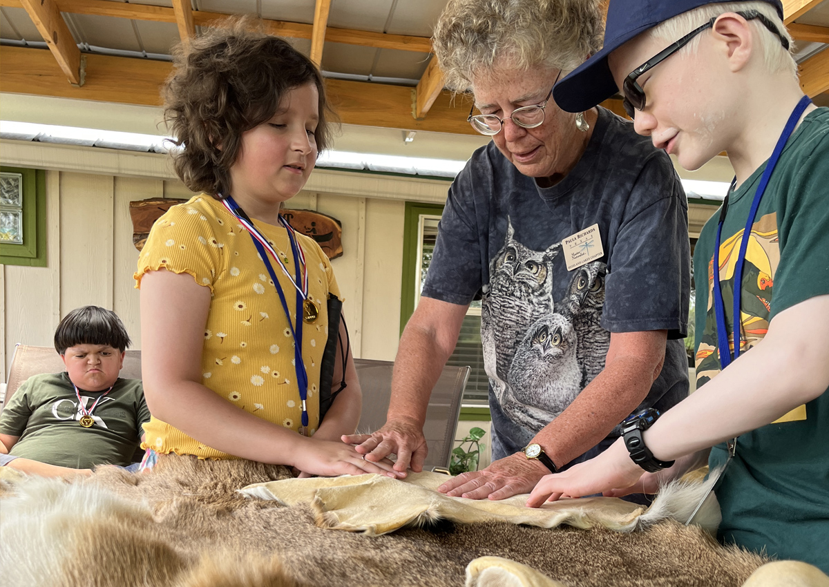 Two adolescents and and adult feel various animal pelts in a cabin. A third adolescent sits in the background.