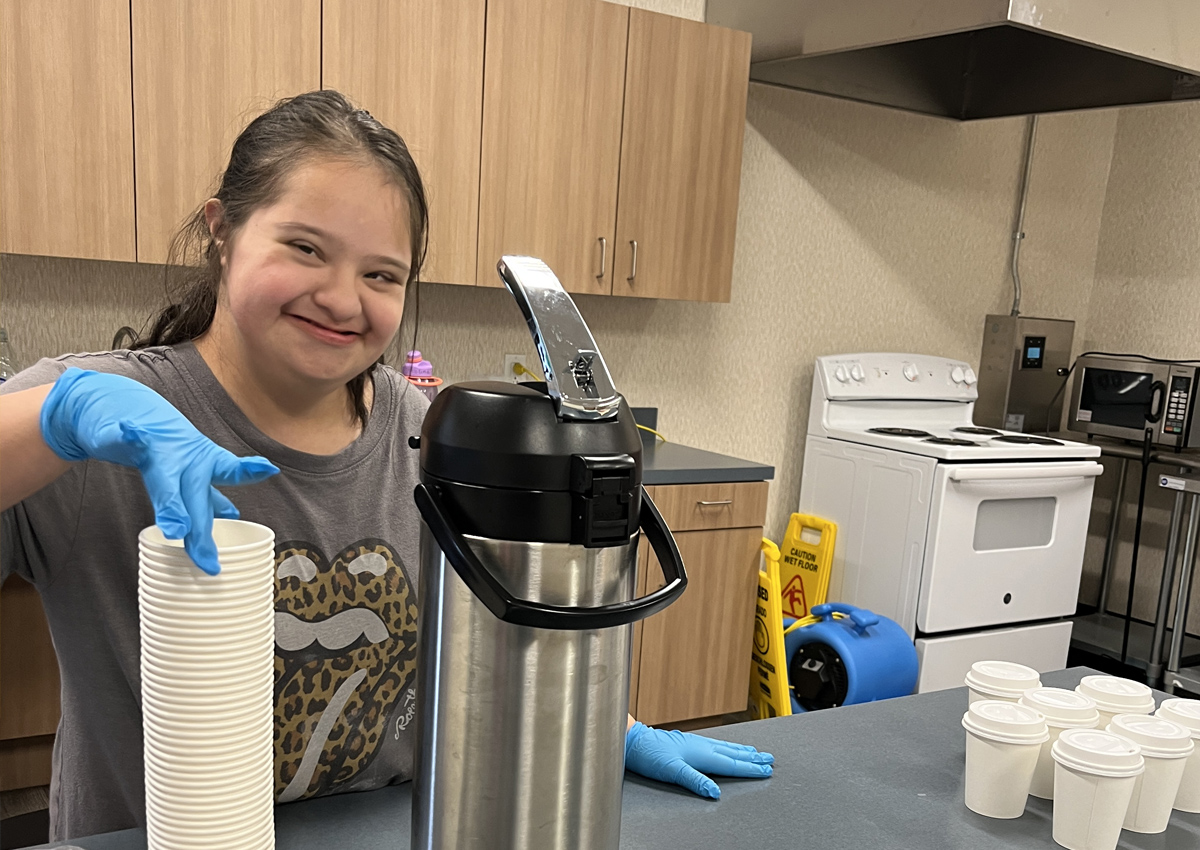 An adolescent takes a paper cup from a stack of paper cups on a counter next to a coffee thermos and cups lined up.